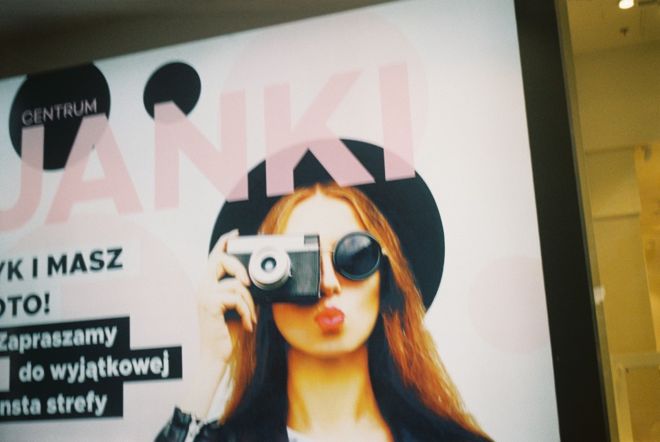 Centrum Janki shopping mall branding advertisement, with a woman in round shades, a black hat and jacket, holding up a Smena 8m to her eye.